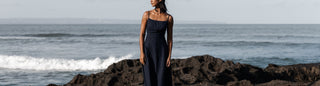 a woman wearing linen loungewear in black coal on the beach with waves crashing on the rocks behind her